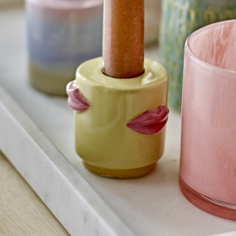 Sassy Stoneware Candle Holder in yellow, a short cylindrical shape with sculpted pink lip details, glossy glaze, holding a metallic brown taper candle, shown beside pastel glassware.