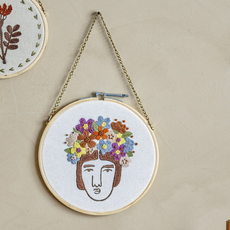Cambiano Cotton Wall Decor in a round wooden hoop, featuring an embroidered face outline crowned with a multicoloured floral wreath on plain cotton, hung on a chain against a neutral wall.