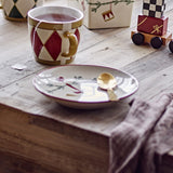 Cream stoneware Christmas side plate with painted burgundy rim and festive branch design, holding a gold spoon, styled on a wooden table beside a tea cup.