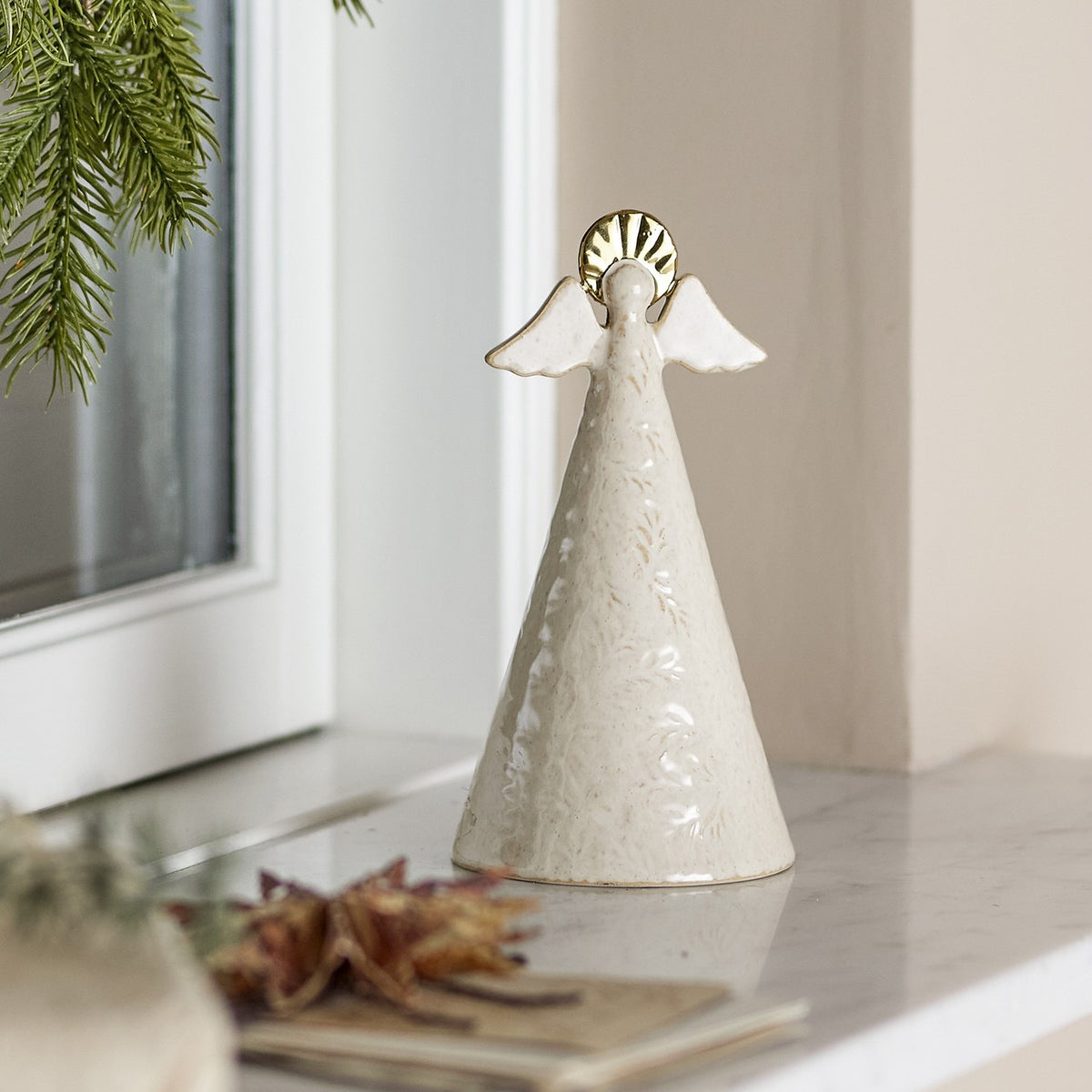Liza Embossed Stoneware Christmas angel decoration in cream, 17cm cone-shaped figure with wings and gold halo, shown on a marble windowsill beside a pine branch.