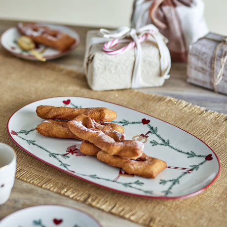 Yule Oval Stoneware Christmas Platter in cream, holding churro-style pastries on a table with wrapped gifts and a matching plate in the background.