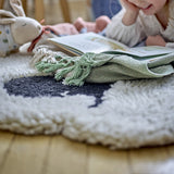 Dolly Sheep Wool Rug in cream with curly pile and dark face detail, shown on a wooden floor beside a plush rabbit toy and folded green throw.