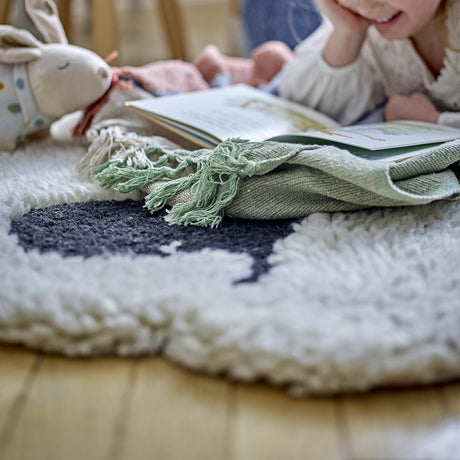 Dolly Sheep Wool Rug in cream with curly pile and dark face detail, shown on a wooden floor beside a plush rabbit toy and folded green throw.