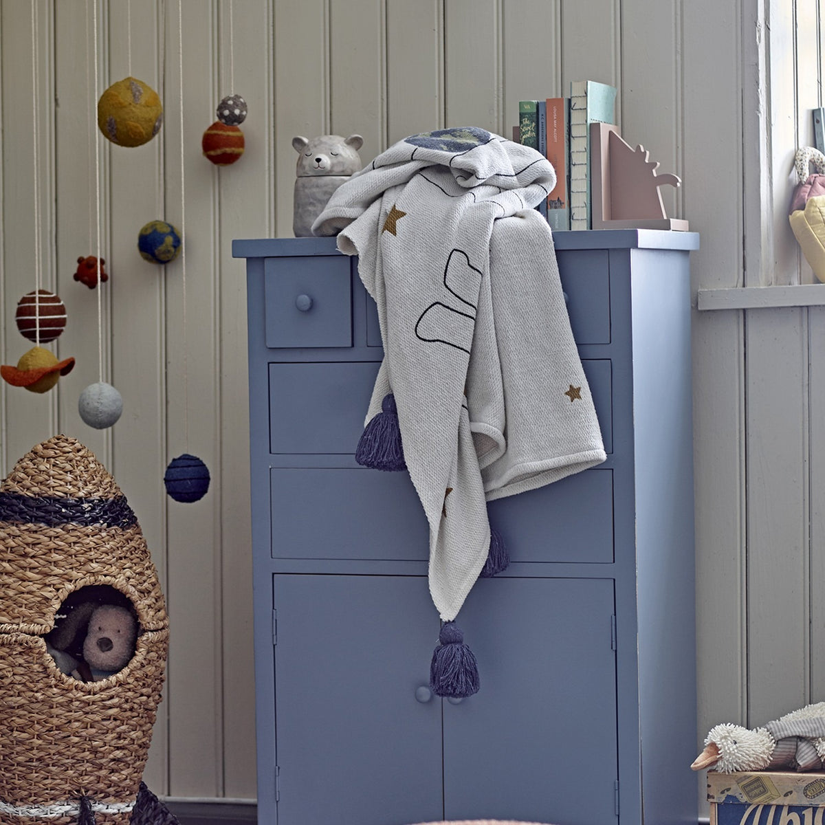 Space Cotton Throw in cream draped over a blue chest of drawers in a nursery, showing a printed rocket, stars and tasselled corners.
