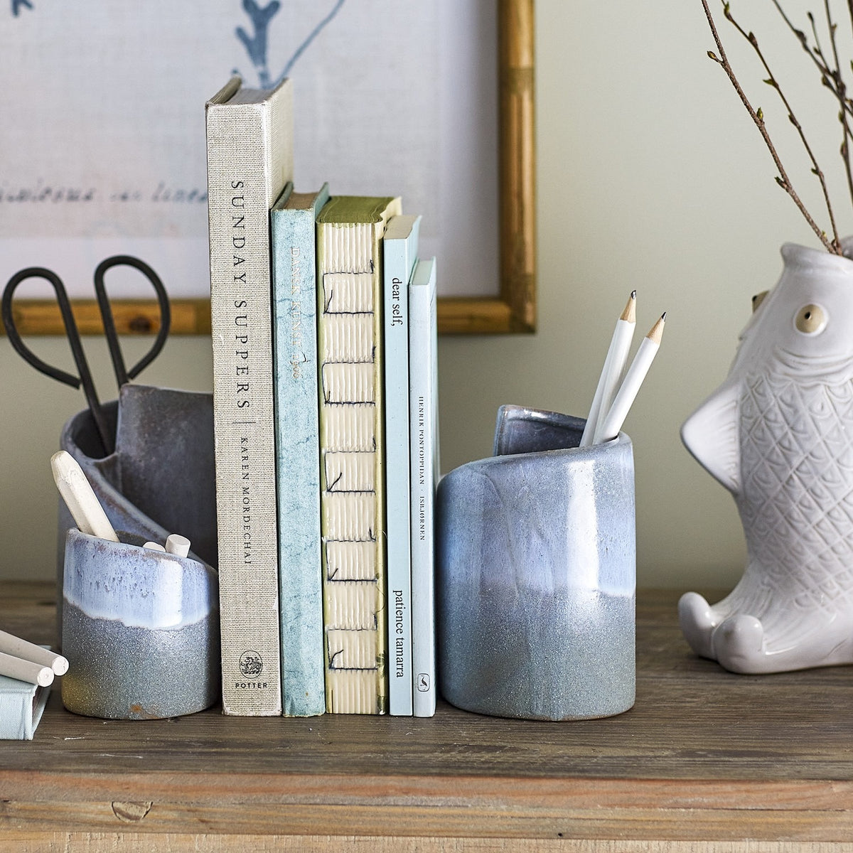 Meliad Stoneware Bookends Set of 2 in blue, curled hollow ceramic forms with glossy light blue rims fading to speckled dark blue bases, shown holding books and pencils on a wooden shelf.