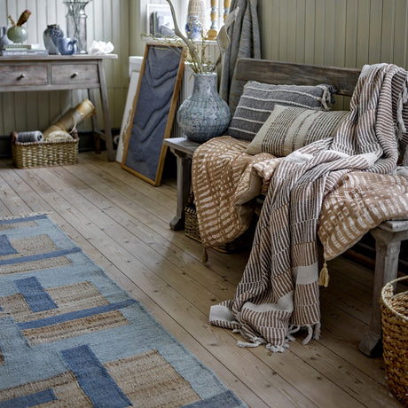 Isnel Brown Striped Cotton Throw with knotted fringe draped over a wooden bench in a rustic living room, layered with striped cushions and a blue woven rug.