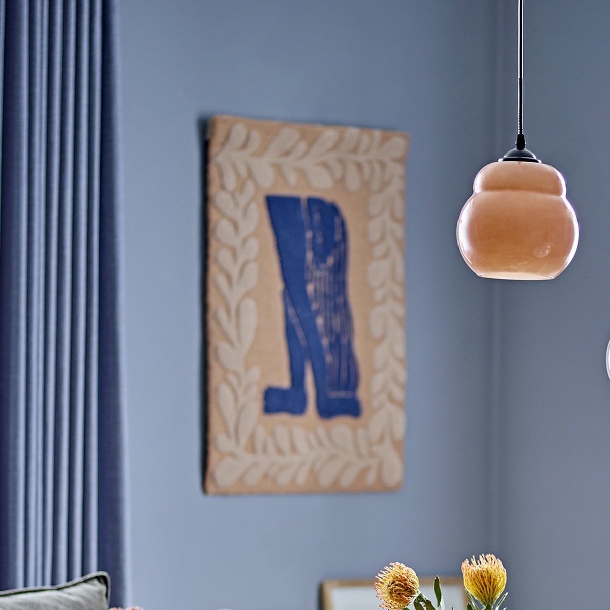 Atle Wool Wall Decor in Multi, a fabric wall hanging with a blue figure motif bordered by leafy sprigs, displayed on a blue wall beside a pendant light.