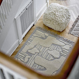 Napoli Wool Rug in grey with raised abstract ivory shapes and fringed ends, shown on a wooden hallway floor beside a shaggy cream pouf.