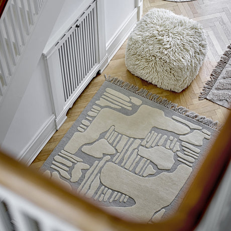 Napoli Wool Rug in grey with raised abstract ivory shapes and fringed ends, shown on a wooden hallway floor beside a shaggy cream pouf.