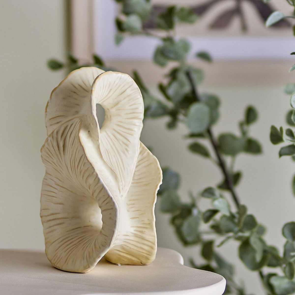 Fabien Stoneware Ornament in cream, a folded ribbon-like sculpture with carved gill-like lines and open centres, shown on a tabletop against a neutral background with greenery.