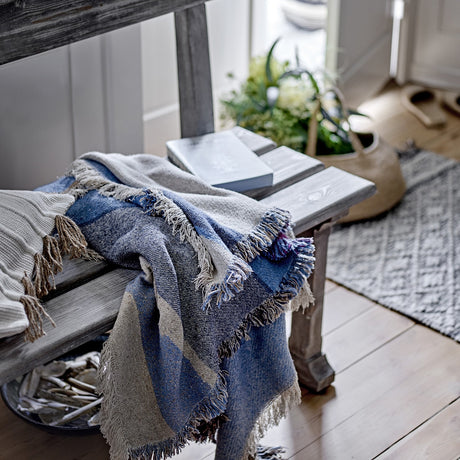 Maji Cotton Throw in multicoloured blue and grey blocks with raw fringed edges, draped over a wooden bench in a rustic hallway setting.