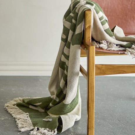 Dalmine Cotton Throw in green, draped over a wooden armchair with leather seat, showing organic printed pattern and knotted fringe edges.