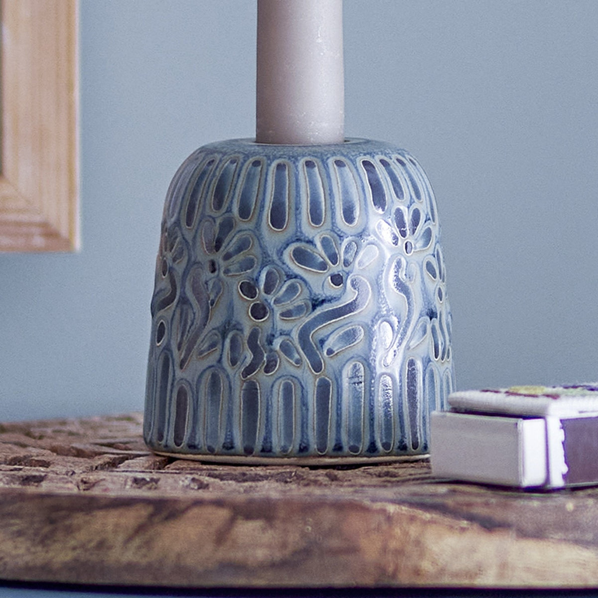 Samsin Floral Stoneware Candle Holder in blue glaze with embossed floral and panel relief, holding a taper candle on a wooden table beside a matchbox.