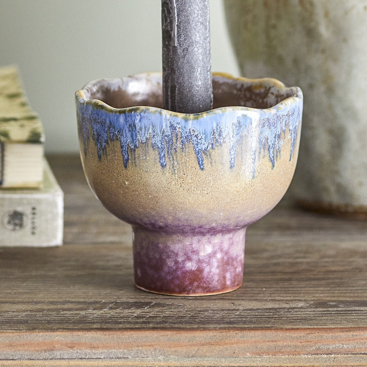 Cloe Stoneware Candle Holder 9cm in Multi, a scalloped-rim pedestal piece with blue drip glaze over a speckled tan body and purple-toned foot, shown holding a candle on a wooden surface.