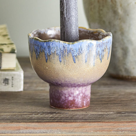 Cloe Stoneware Candle Holder 9cm in Multi, a scalloped-rim pedestal piece with blue drip glaze over a speckled tan body and purple-toned foot, shown holding a candle on a wooden surface.