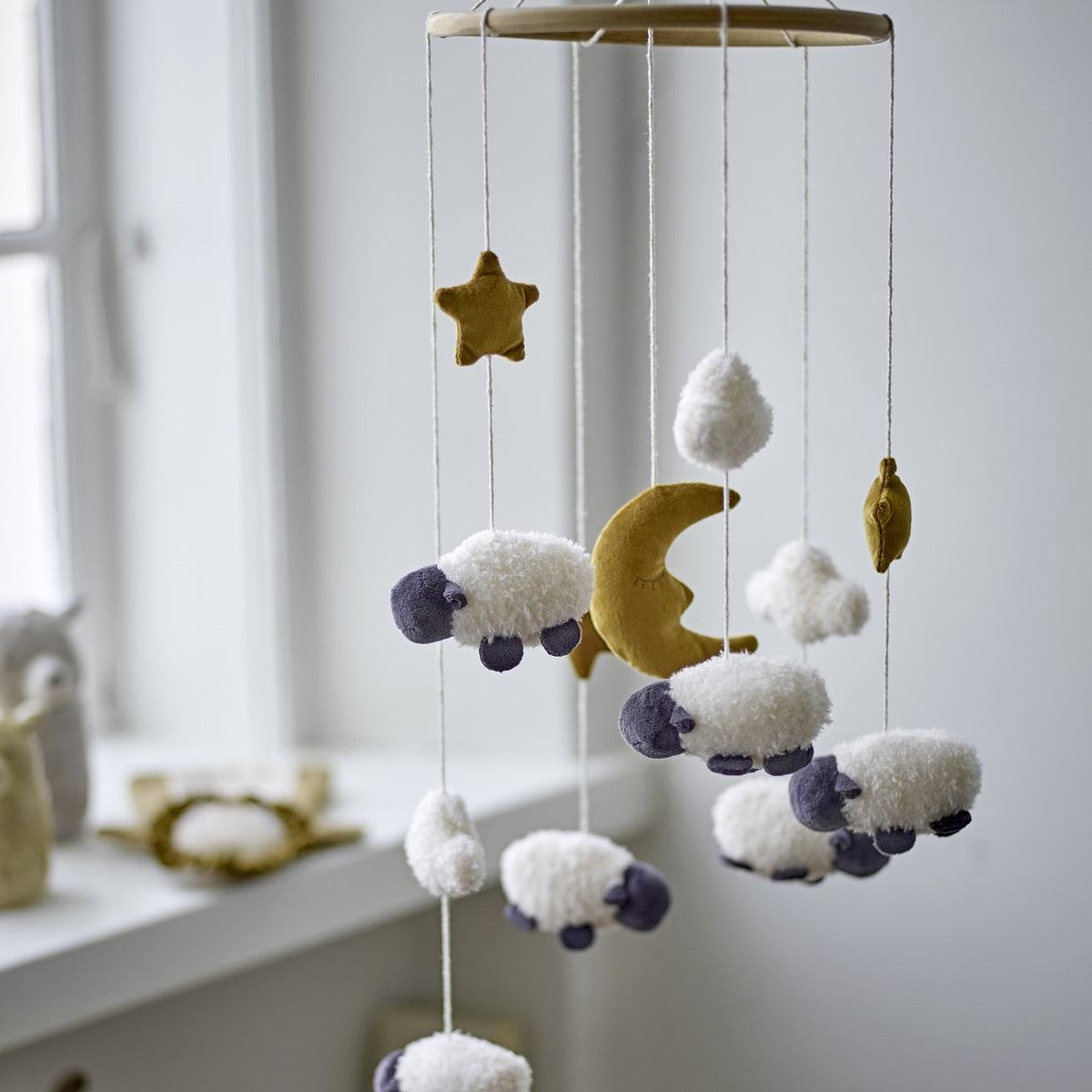Dolly Star Baby Mobile in white, a round wooden hoop with hanging fabric sheep, crescent moon, star and cloud shapes, shown in a nursery setting near a window.