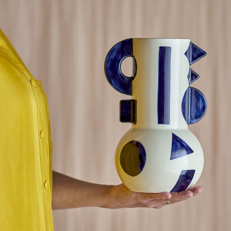 Graphica Geometric Stoneware Vase 24cm in cream, held in hand, showing its tall neck with hand-painted blue stripes, triangles and circles plus raised D-shaped, square and triangular details.