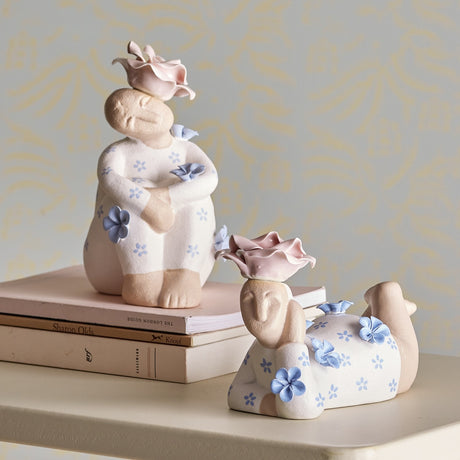 Floriella Floral Stoneware Ornaments Set of 2 - two seated and reclining ceramic figurines with floral patterns and sculpted rose hats, displayed on books against a patterned wall.