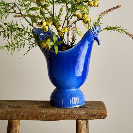 Wylie Bird Stoneware Vase 29cm in Blue, with a crested head and curved beak, ribbed fanned rim, and glossy streaked glaze, shown on a wooden stool holding greenery and berries.