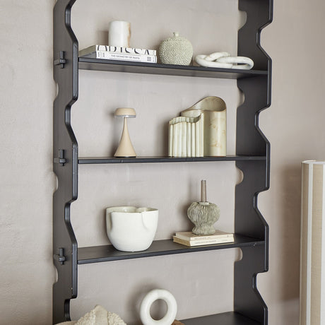 Savoya wooden wall shelf in black, tall fir shelving unit with wavy stepped side panels, displaying books, vases, a lamp and ceramic decor against a neutral wall.