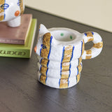 Caty Striped Stoneware Candle Holder in multicoloured ceramic, cat-shaped with a ribbed stacked body and looped tail handle, shown on a dark surface beside books.