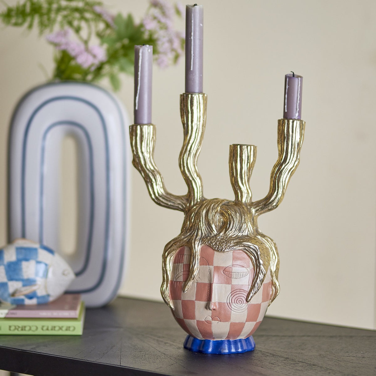 Medusa Resin Candle Holder in gold with a checked face, gold tendril crown, and four branching arms holding lilac candles, styled on a shelf with books and a vase.