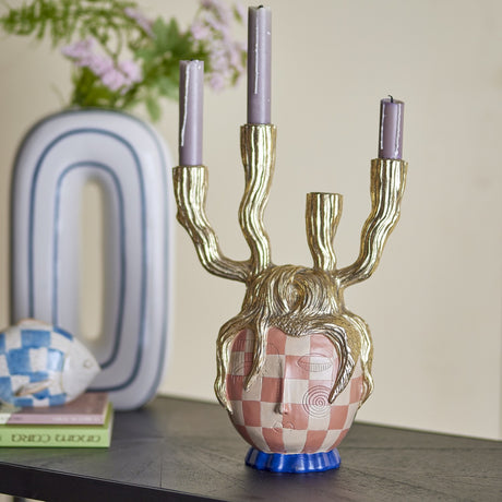 Medusa Resin Candle Holder in gold with a checked face, gold tendril crown, and four branching arms holding lilac candles, styled on a shelf with books and a vase.
