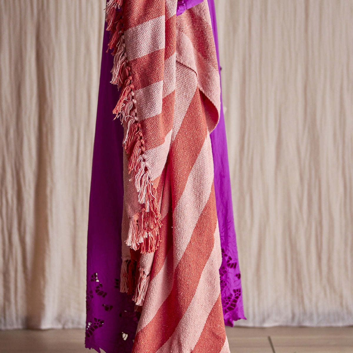 Frello Striped Cotton Throw in orange and pink tones, draped over a purple garment, showing diagonal stripes and a knotted fringe edge.