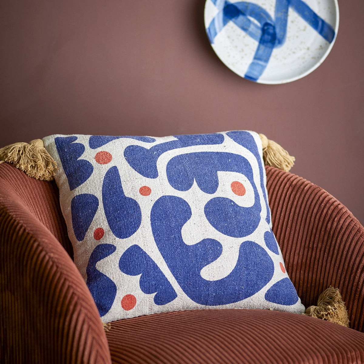 Eviaya Cotton Cushion in Multi, square with tasselled corners, printed with bold blue abstract shapes and orange dots, shown on a rust corduroy armchair.