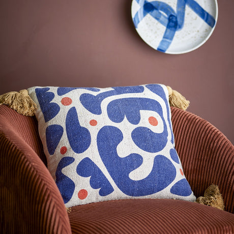 Eviaya Cotton Cushion in Multi, square with tasselled corners, printed with bold blue abstract shapes and orange dots, shown on a rust corduroy armchair.