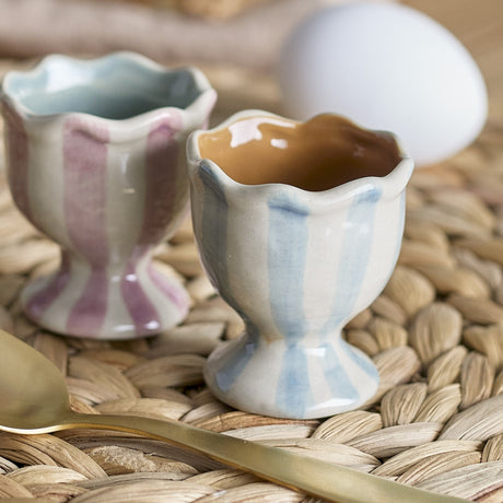 Willa Striped Stoneware Egg Cups, set of 2, with wavy rims and pink and blue vertical stripes on a round footed base, shown with an egg and spoon on a woven mat.