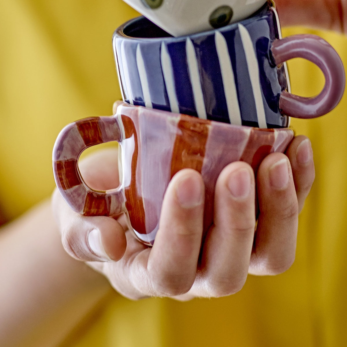 A hand holds the Nini Brown Striped Stoneware Cup with white and navy vertical stripes and a purple handle, stacked atop a pink striped ceramic mug.