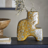 Ollia Stoneware Vase 20cm in Brown, a sculptural stepped ceramic vase with mottled speckled glaze, arched base and two feet, shown with dried branches on a dark console beside a wooden bowl and books.