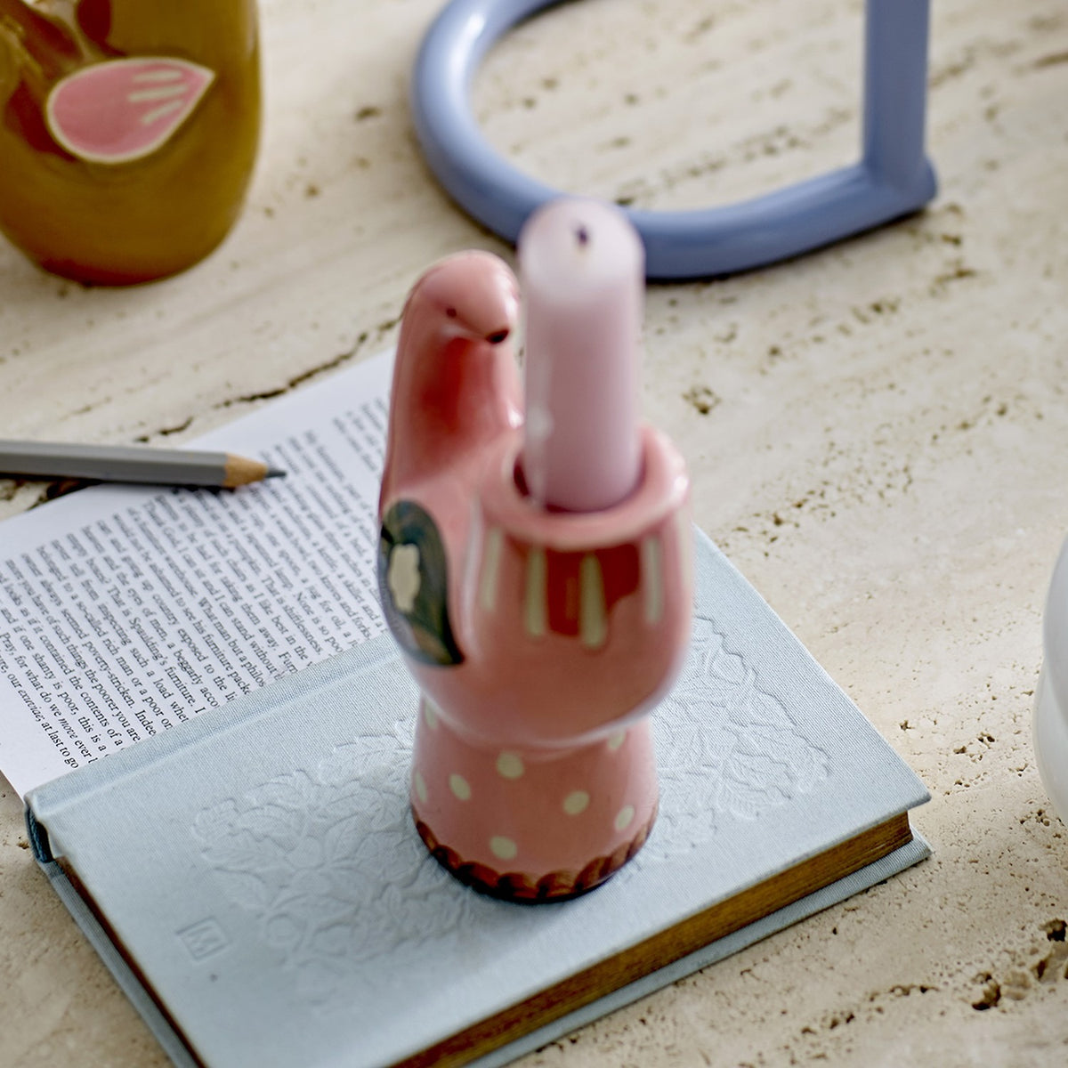 Trudy Bird Stoneware Candle Holder in pink ceramic, shaped like a bird with a polka-dot base, holding a single taper candle, displayed on a book beside a pencil and open notebook.