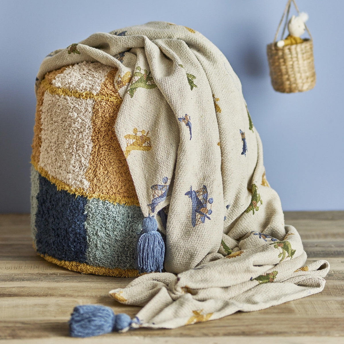 Plane Cotton Throw in beige, patterned with biplanes and clouds, blue tassel corners, draped over a tufted ottoman beside a woven basket decoration.