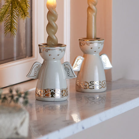 Faella Angel Christmas Candle Holders, Set of 2, white ceramic figures with gold scrolled hems and wings, holding twisted candles on a marble shelf.