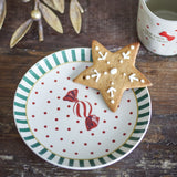 Yule Striped Stoneware Christmas Plate in Multi, a small 16cm side plate with a green striped rim, red polka dots and a wrapped sweet design, shown with a star-shaped cookie on a wooden surface.