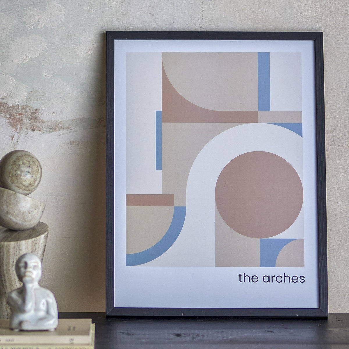 Arches Geometric Wooden Framed Print in black frame, featuring overlapping curves, arches and a circle in soft tan, blush and blue tones, leaning against a wall beside decorative objects.