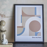 Arches Geometric Wooden Framed Print in black frame, featuring overlapping curves, arches and a circle in soft tan, blush and blue tones, leaning against a wall beside decorative objects.