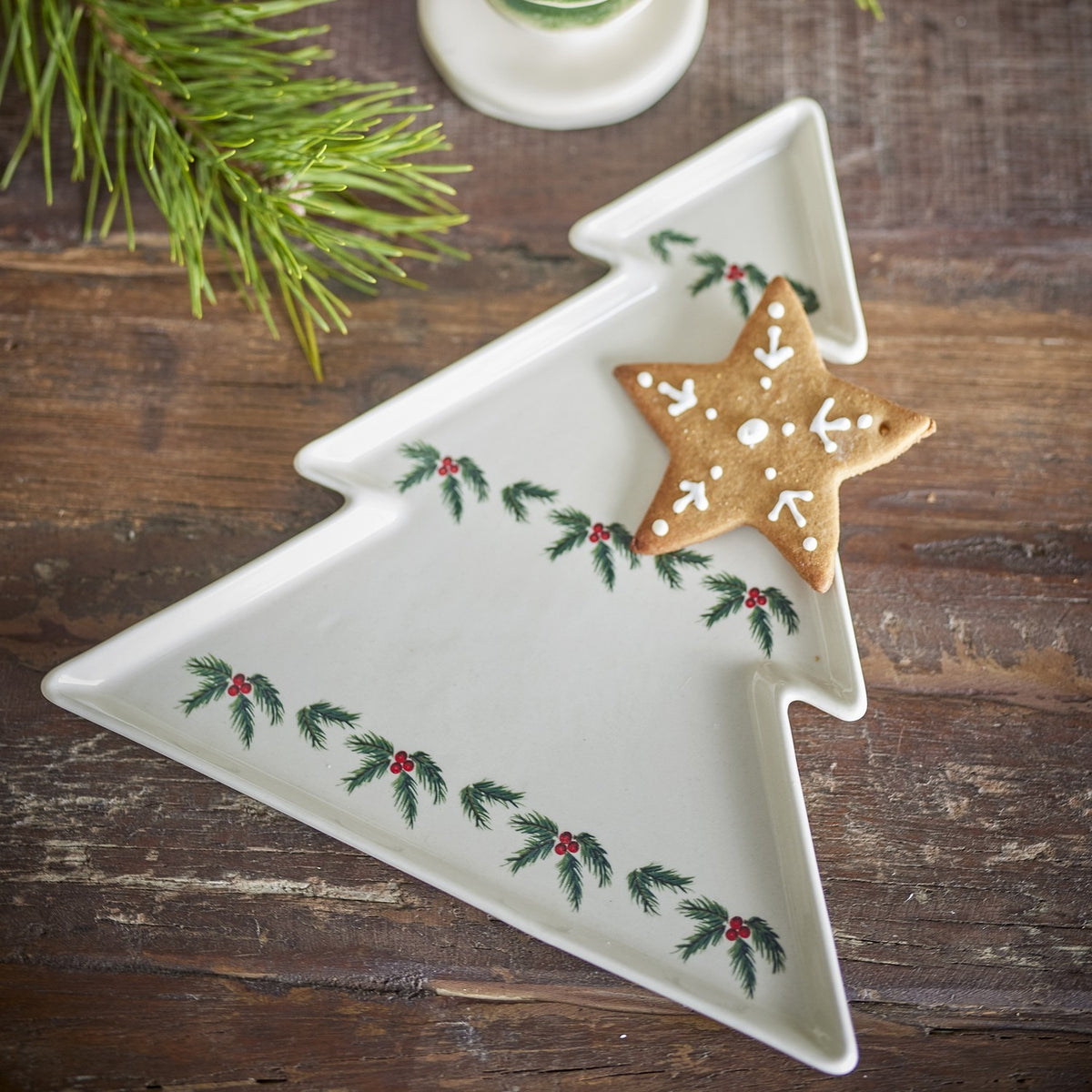 Berry stoneware Christmas platter in white, tree-shaped with stepped tiers and low rim, decorated with painted holly sprigs and berries, shown with a star-shaped cookie on a wooden table.