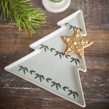 Berry stoneware Christmas platter in white, tree-shaped with stepped tiers and low rim, decorated with painted holly sprigs and berries, shown with a star-shaped cookie on a wooden table.