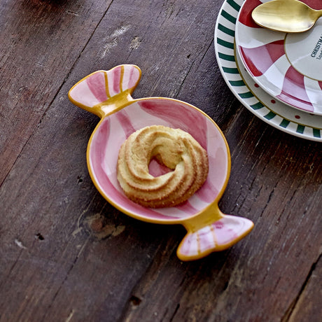 Caramella Wrapped Sweet stoneware Christmas platter in pink, shaped like a twisted candy wrapper with hand-painted swirls, shown holding a swirled shortbread biscuit on a wooden table.