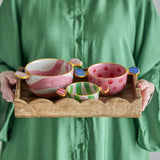 Caramella sweet-shaped ceramic bowls, set of three in pink swirl, pink polka-dot, and green stripe patterns with twisted wrapper handles, held on a wooden tray by a person in a green shirt.