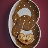 Stoneware serving platter shaped like a pretzel, glossy brown glaze with white dot detailing, shown with pastry swirls on a dark red background.