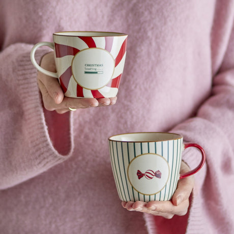 Two stoneware Christmas mugs held in hands against a pink jumper, one with a red candy-swirl pattern and "Christmas loading" text, the other with teal stripes and a sweet motif.