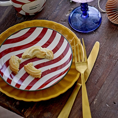 Jolly Striped Stoneware Christmas Plate in red and white swirl pattern, shown on a mustard yellow plate with shortbread biscuits, gold cutlery and a blue glass goblet on a wooden table.