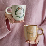 Jolina stoneware Christmas mugs held in hands, one cream with green wreath and "Merry Christmas" text, one beige with snowflakes and "let it snow" text.