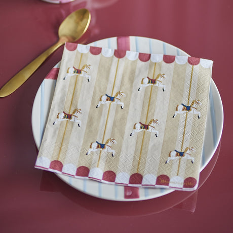 Folded paper napkin from the Merry-go-round pack of 20, printed with carousel horses on striped cream and scalloped red-and-white edging, set on a white plate beside a gold spoon.