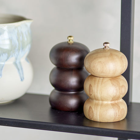Marge Wooden Salt & Pepper Grinders Set of 2 in dark and light wood, each with stacked rounded tiers and a brass-tone knob, shown on a dark shelf beside a jug.
