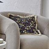 Eydis Floral Cushion in Multi, a fringed square cushion with dense floral print in gold, burgundy and white on black, shown on a beige armchair.